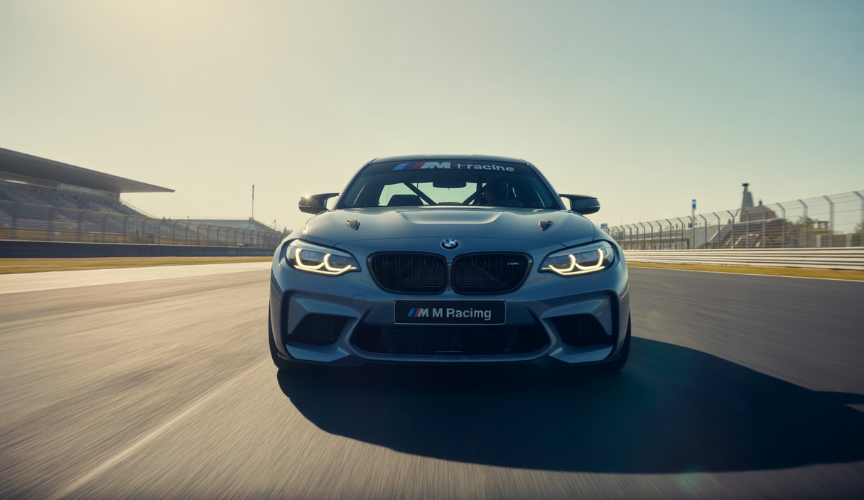 BMW M2 Racing vue de face sur circuit, éclairage adaptatif LED avec anneaux jaunes, design sportif aérodynamique et carrosserie noire avec éléments en fibre de carbone, ambiance dynamique de course en journée ensoleillée