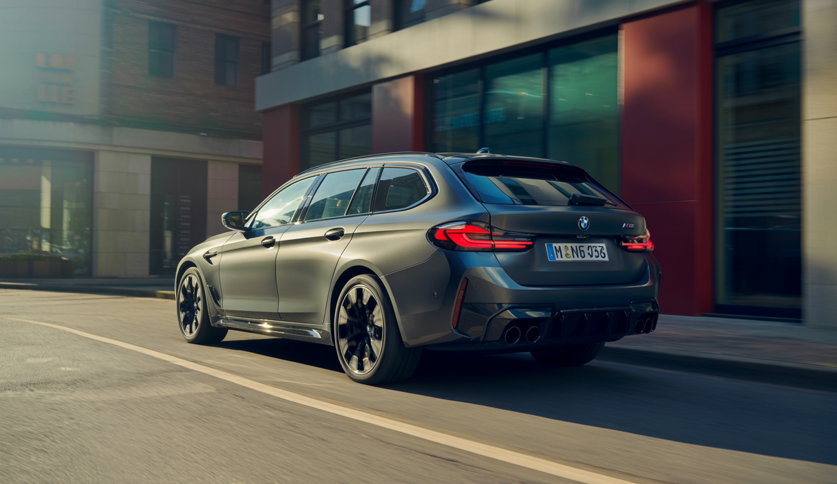 BMW M5 G99 Touring 2025 vue avant latérale dans un cadre urbain moderne, break sportif hybride avec design distinctif M, grande surface vitrée, peinture métallisée sombre et jantes alliage larges